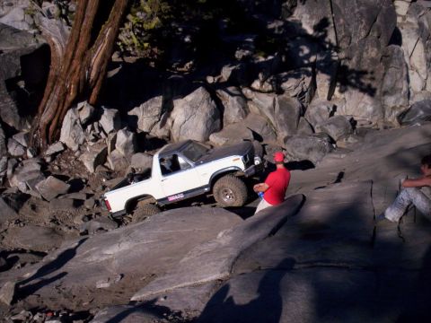 1989 Ford Bronco - A white and grey pickup truck is shown off-roading on a rocky slope with a person in a red shirt observing.