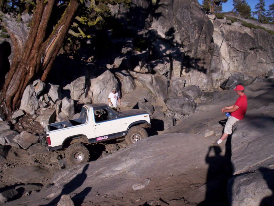 White off-road SUV with a black top driving over rocks, a person in a red shirt sits on a rock nearby