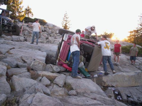 1989 Ford Bronco - A group of people are gathered around a red Jeep Wrangler that has rolled onto its side on a rocky incline.