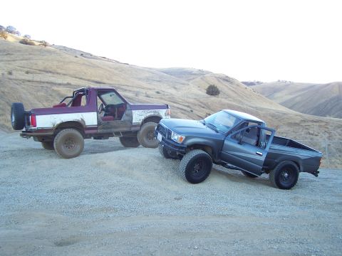 1991 Ford Bronco - Two off-road vehicles, a Ford Bronco and a Toyota pickup, are parked on a dirt track with hills in the background.
