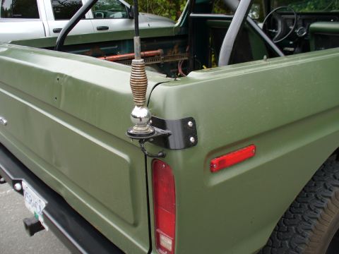 1978 Ford Bronco - Close-up of the rear driver-side corner of an olive/sage green open-top 4x4 with the top removed, showing a whip CB antenna on a black brac…