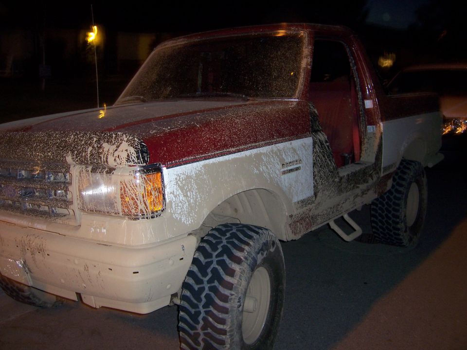 Mud-covered red and white Ford Bronco with off-road tires, driver's door open