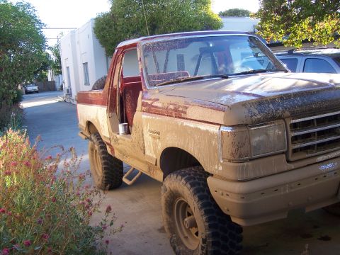 1991 Ford Bronco - A muddy red and white Ford Bronco is parked outside, with another vehicle visible in the background.