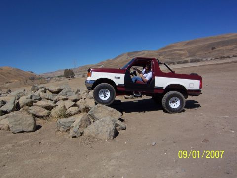 1991 Ford Bronco - A maroon and white 1991 Ford Bronco navigates a rocky terrain under a clear blue sky.