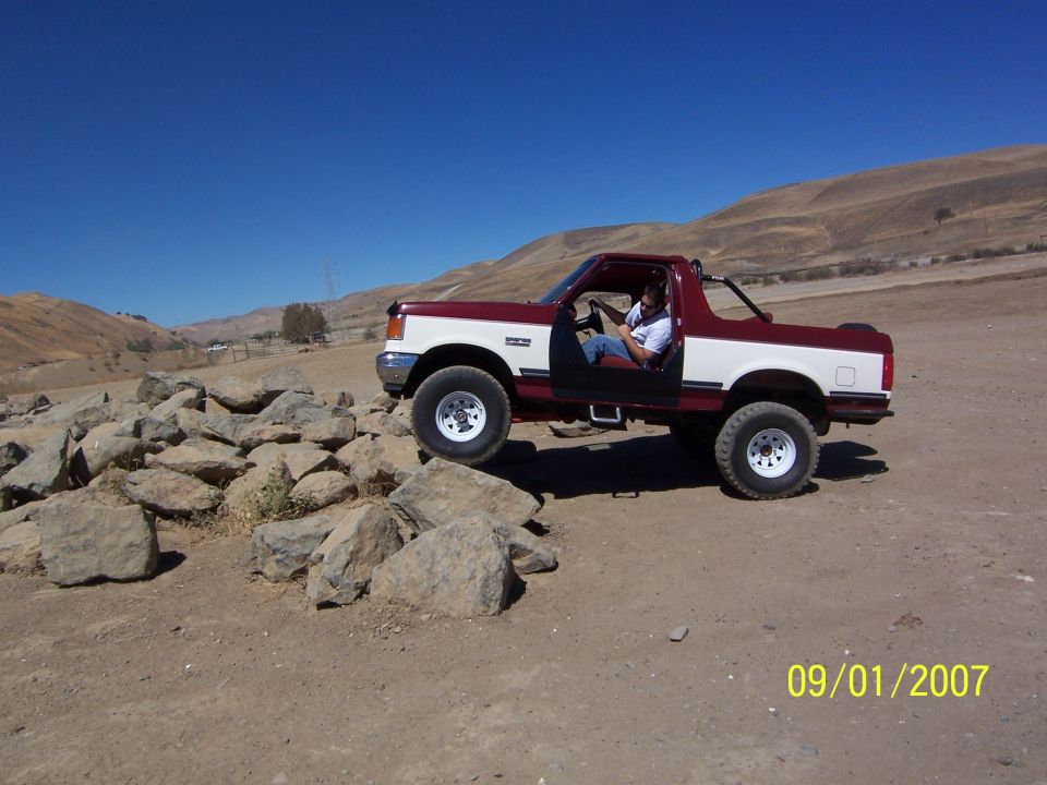 Maroon and white 1991 Ford Bronco with doors removed, driving over rocks