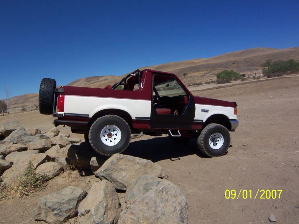 Maroon and white Ford Bronco on rocks, spare tire on back. Doors are off
