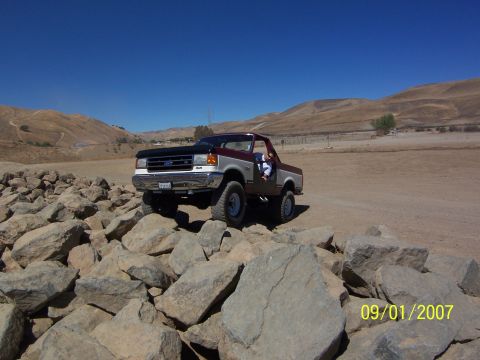 1991 Ford Bronco - A two-tone maroon and silver Ford F-Series truck navigates a rocky terrain under a clear blue sky.