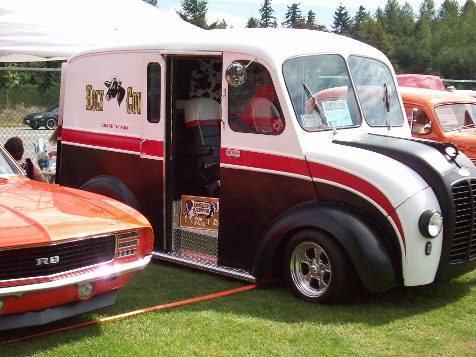 Vintage delivery truck with 'Holy Cow' logo and an orange muscle car next to it