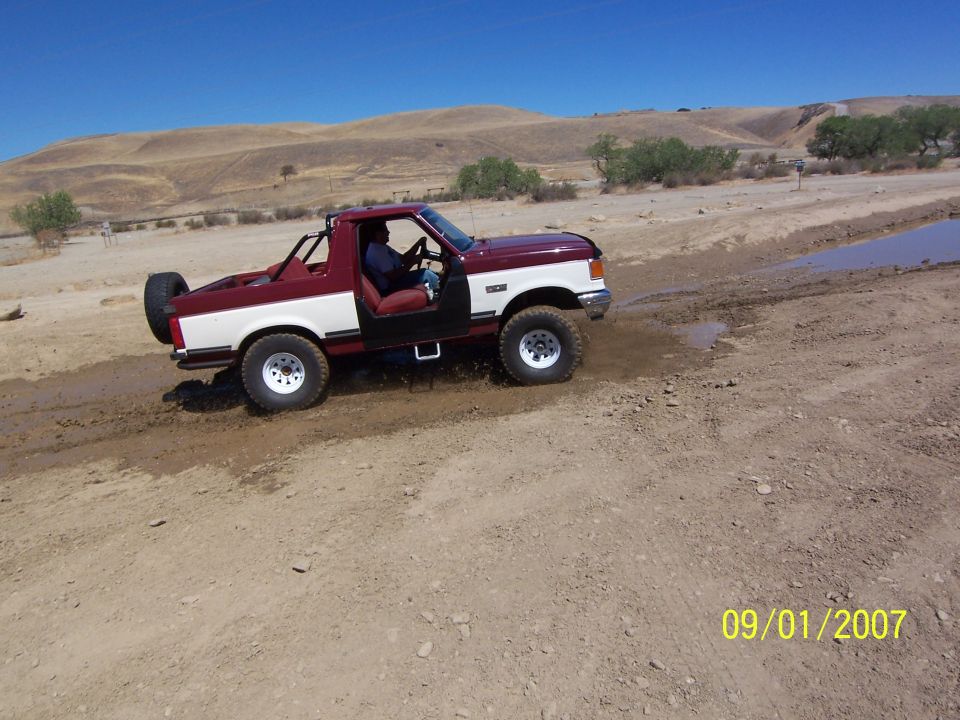 Maroon and white Ford Bronco with doors removed, driving through mud