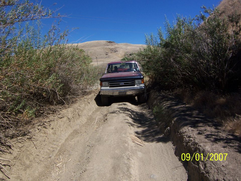 1991 Ford F-Series truck driving on a rutted dirt road between brushy hills