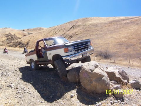 1991 Ford Bronco - A two-tone Ford F-Series truck navigates a rocky trail with two dirt bikes in the background.