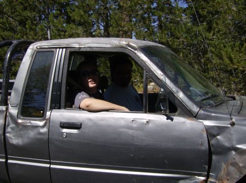 1986 Ford Bronco - A silver pickup truck with significant body damage is seen from the side, with two people inside the cab.