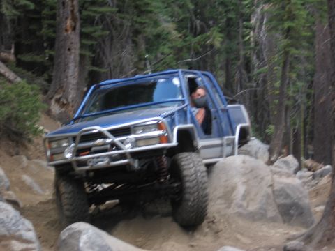 1994 Ford Bronco - Off-road pickup truck with custom roll cage climbs over rocks on a forest trail.