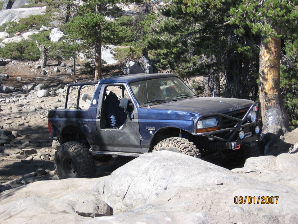 Blue topless Ford Bronco with roll bar and winch crawling granite boulders in pines