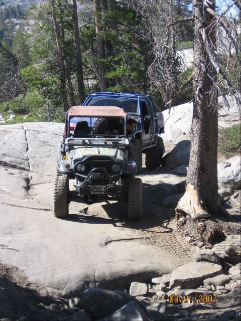 1994 Ford Bronco - Two heavily modified off-road vehicles traverse a challenging rocky terrain on a sunny day.