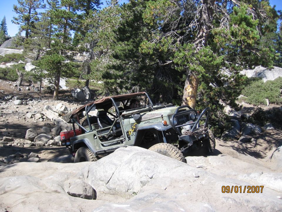 Green doorless Jeep with roll cage and winch crawling off a granite slab in pine forest