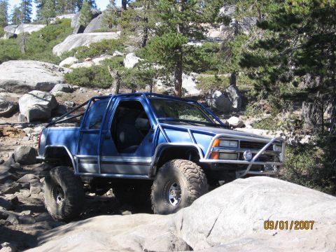1994 Ford Bronco - This modified pickup truck is shown off-roading on a rocky trail with trees and boulders in the background.