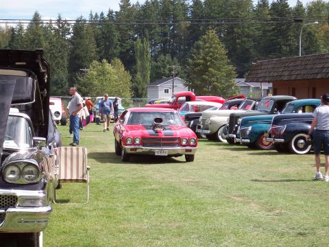 1997 Ford Mustang - A red muscle car with a large hood scoop and racing stripes is displayed at an outdoor car show alongside other vintage vehicles.
