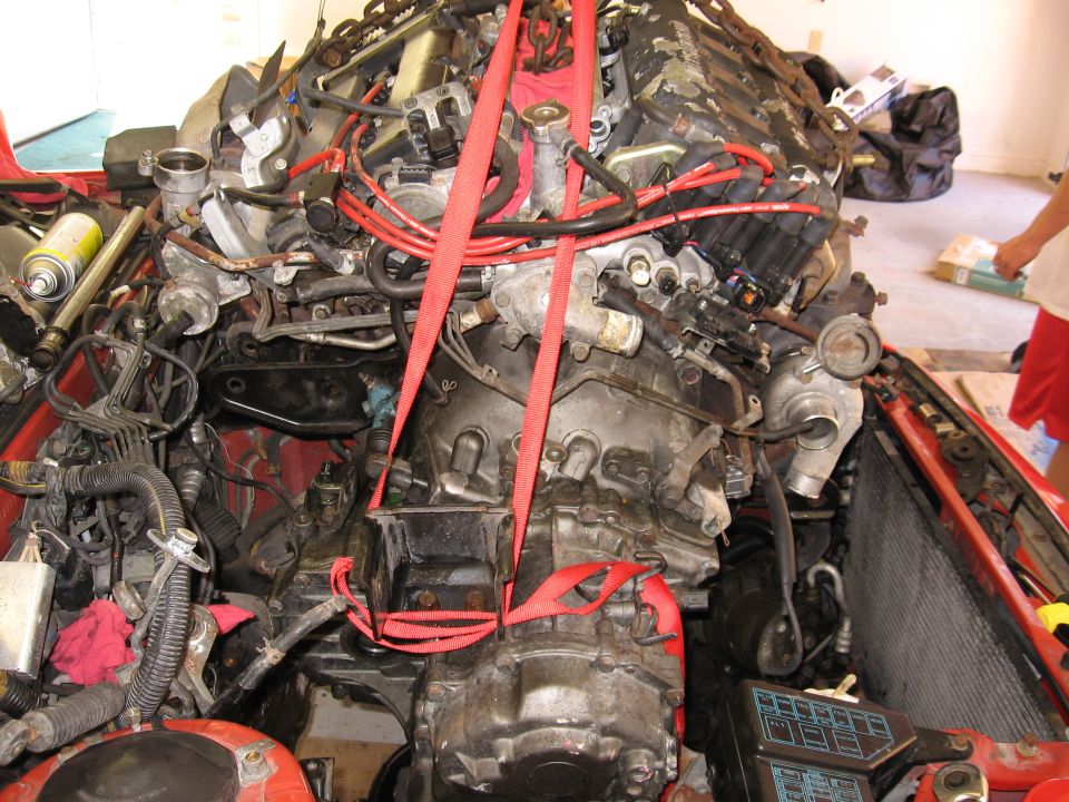 Engine being lifted out of a red car with red straps. Wires and hoses are visible
