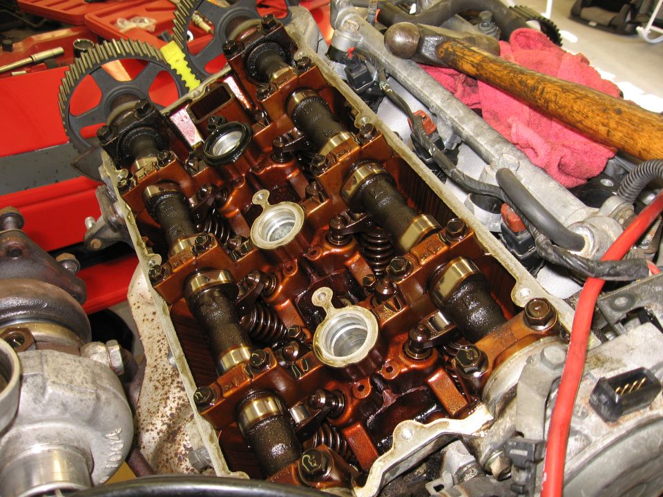 Engine cylinder head with camshafts, valves, and springs visible. Tools are nearby