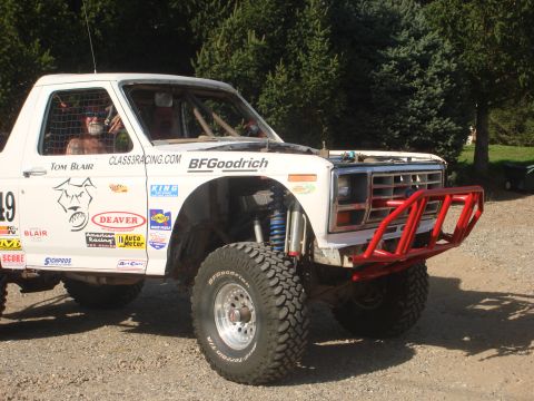 1990 Ford Bronco - white number 49 off-road race truck with BFGoodrich tires, red tube bumper and sponsor decals parked on gravel.