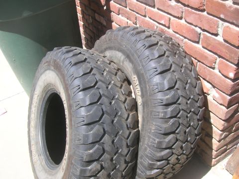 1985 Ford Bronco - A close-up view of two off-road tires, one with a white sidewall, resting against a brick wall.
