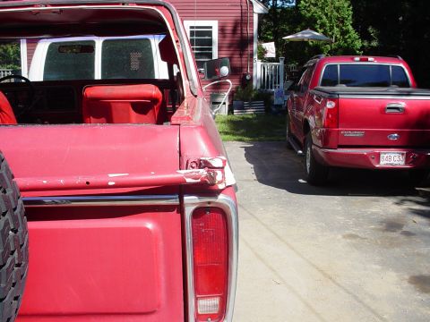 1978 Ford Bronco - Close-up of the rear of a red pickup truck, showing damage to the tailgate and a spare tire. A second red SUV is visible in the background.