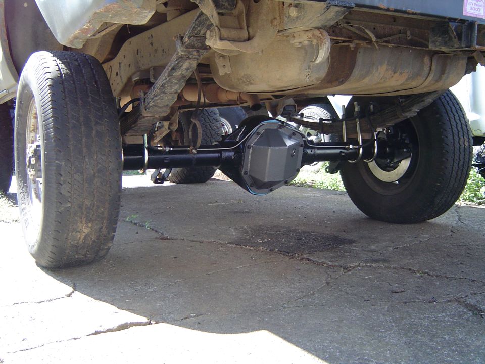 Underside view of a vehicle's rear axle, leaf springs, and fuel tank. Another vehicle is partially visible behind it
