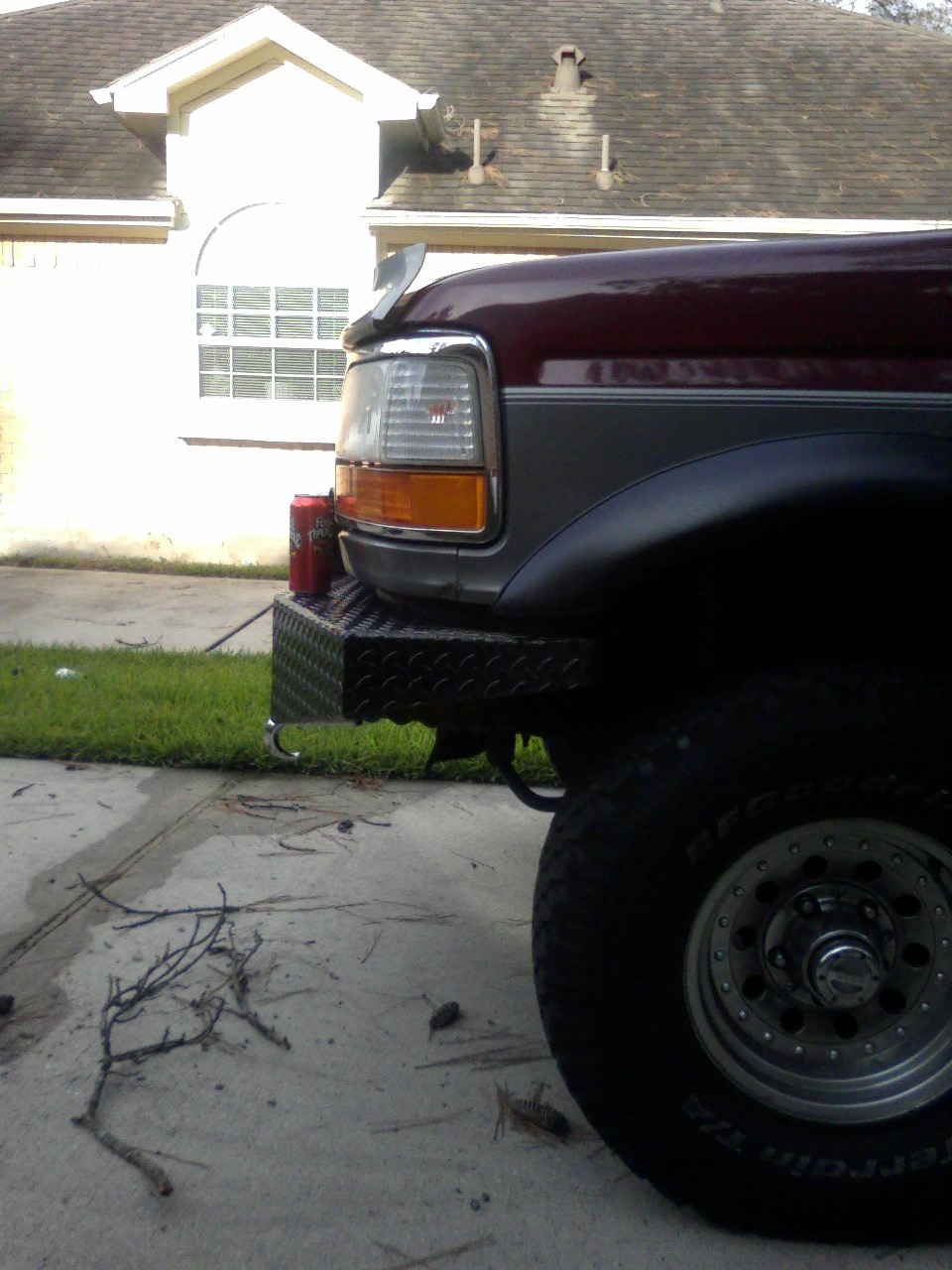 Front corner of lifted burgundy Ford Bronco with diamond-plate bumper and BFGoodrich A/T tire in driveway