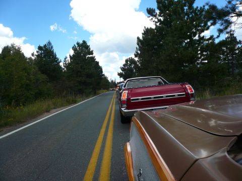 1975 Ford Ranchero - A red 1975 Ford Ranchero is seen from behind, driving in a line of vehicles on a rural road.