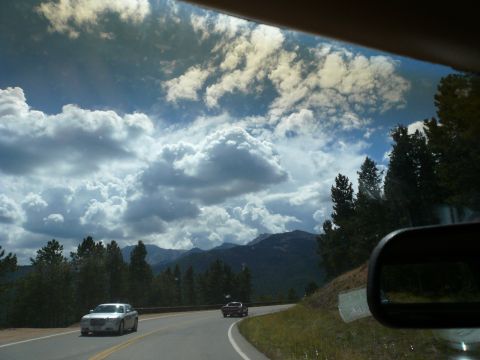 1975 Ford Ranchero - Two cars travel along a scenic mountain highway, with dramatic clouds overhead and pine trees lining the roadside.