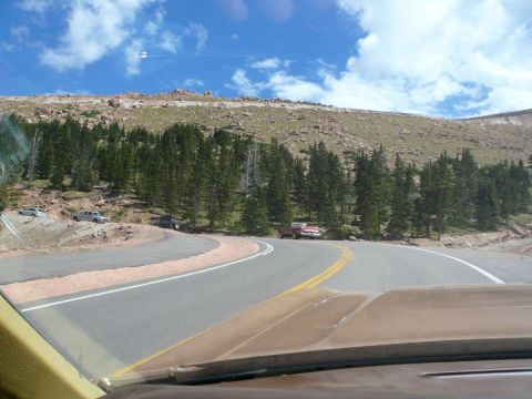 1975 Ford Ranchero - Several vehicles are parked along a winding mountain road with trees and rocky hillsides under a cloudy sky.