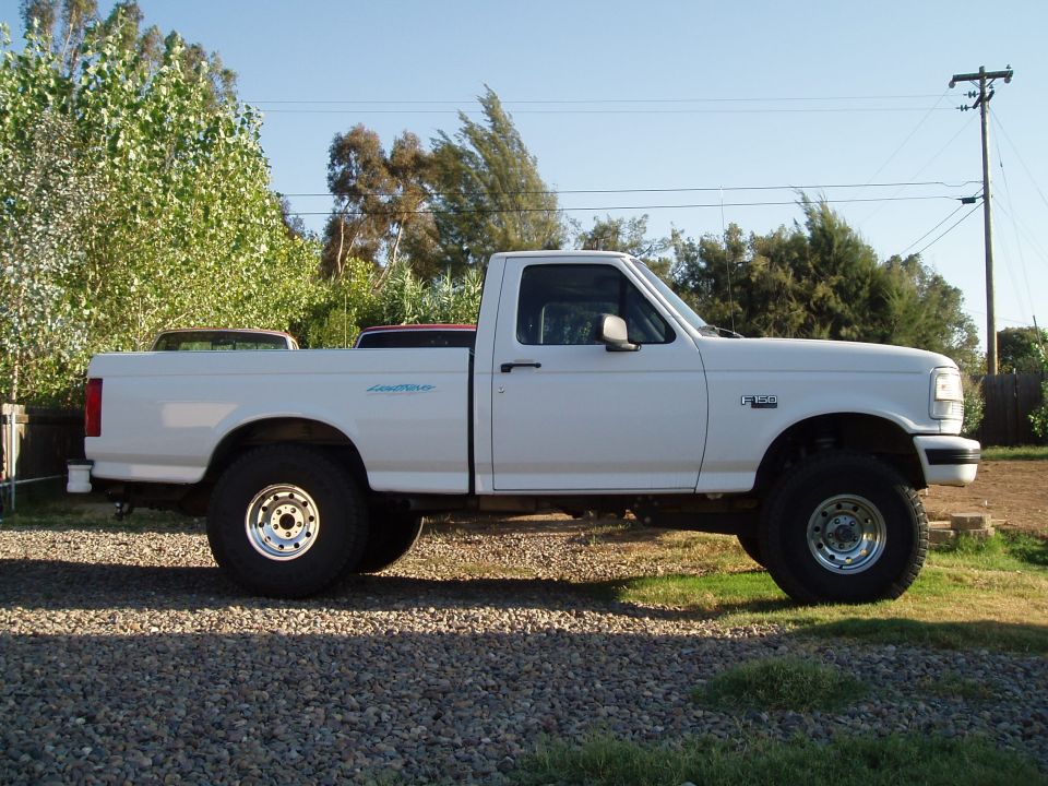 White 1994 Ford F-150 pickup truck, side view, lifted suspension, chrome wheels, parked on gravel