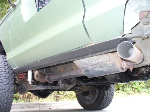 1978 Ford Bronco - Low-angle view along the passenger side of a lifted sage green 4x4, showing a black rock slider/rocker guard, an angled exhaust tip exiting…