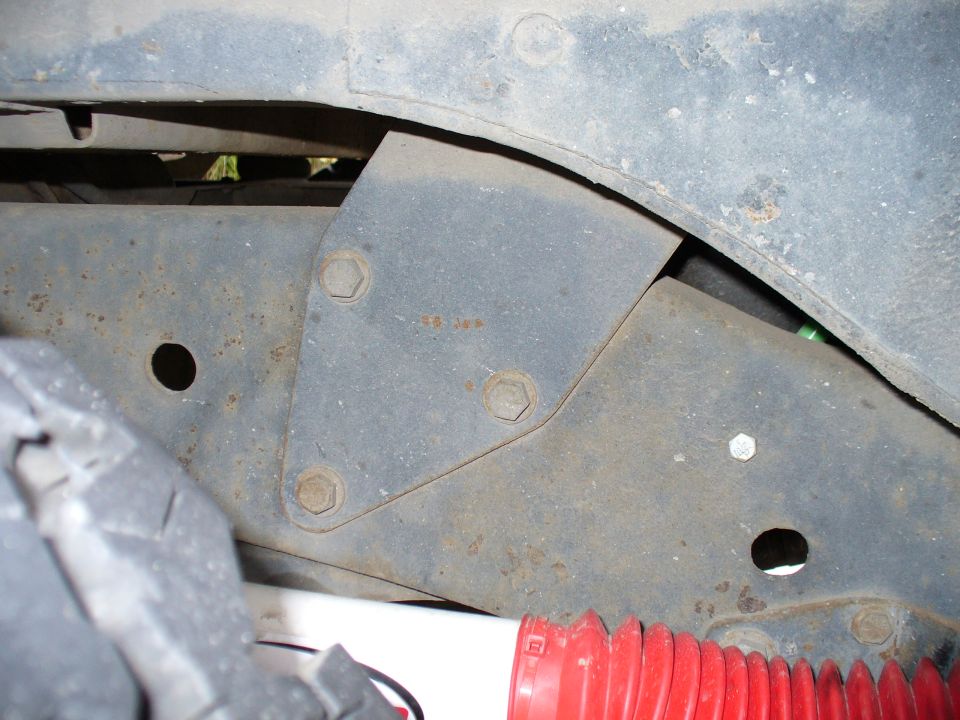 Metal bracket with three bolts attached to a vehicle frame, near a tire and red shock absorber