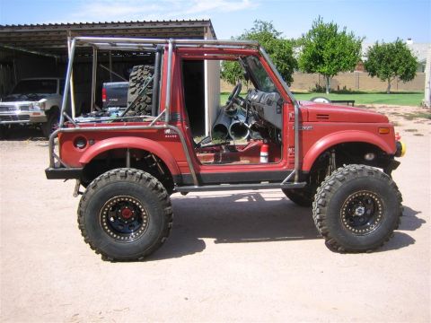 1987 Suzuki Samurai - A modified red 1987 Suzuki Samurai with an external roll cage and off-road tires sits outside a garage.