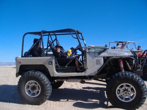 1986 Ford Bronco - Two off-road vehicles are parked on a sandy surface with a clear blue sky overhead.