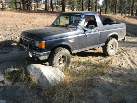 1988 Ford Bronco - This 1988 Ford Bronco is seen off-road, parked on a dirt path with trees and a fence in the background.