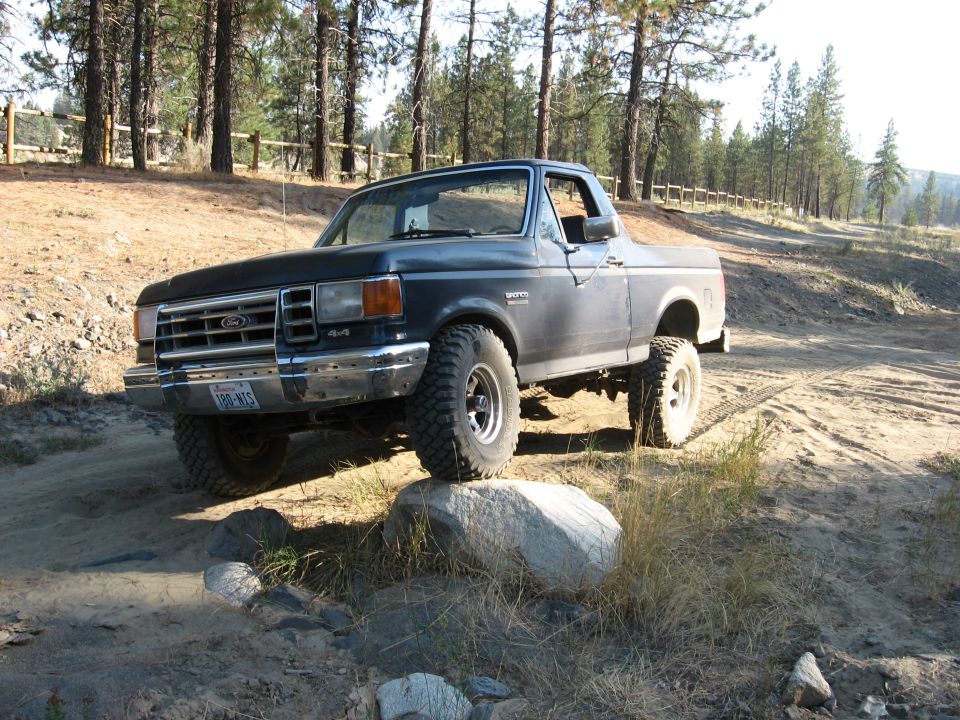 Dark blue Ford Bronco on a dirt trail, front tire on a rock. Trees and a wooden fence in background