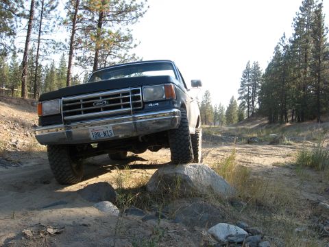 1988 Ford Bronco - A black Ford truck navigates a rocky, off-road trail surrounded by trees.
