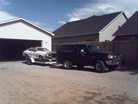 1988 Ford Bronco - A black Ford Bronco tows a white car on a trailer near a residential garage.