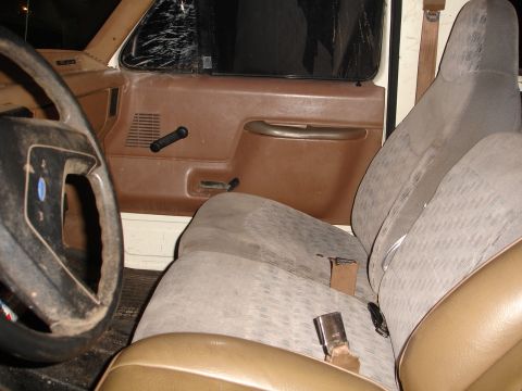 1989 Ford Bronco - The interior of a vehicle shows brown door panels and worn cloth seats, with a cracked windshield visible.