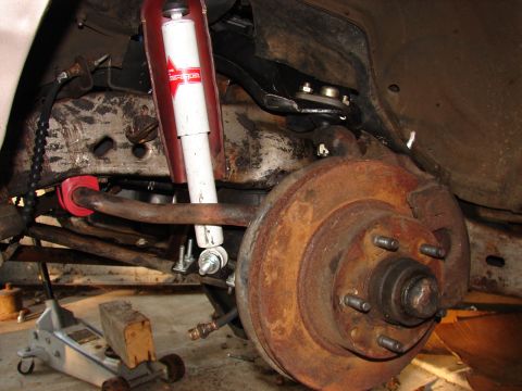 1973 Ford Ranchero - Close-up of a front suspension and brake assembly with a rusty disc rotor and five-lug hub, a new white gas shock absorber with red upper b…