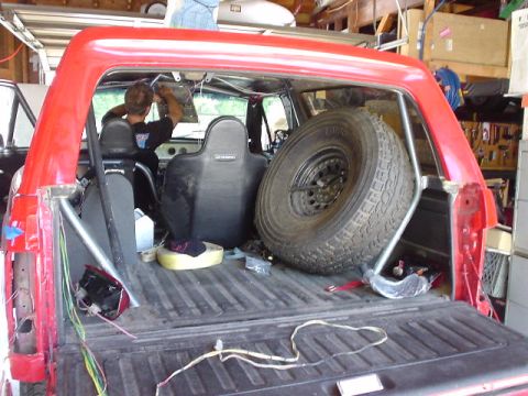1984 Ford Bronco - The interior of a red Ford Bronco shows a roll cage, spare tire, and exposed wiring, with a person working on the dashboard.