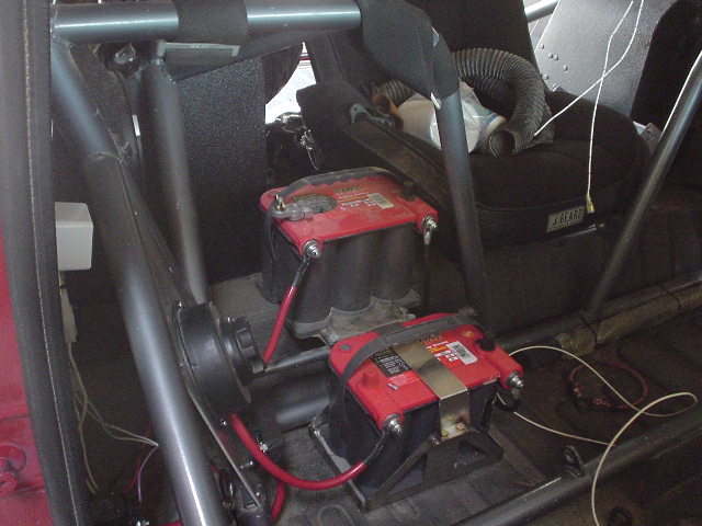 Two relocated red-top batteries mounted behind seats inside a caged, stripped race vehicle