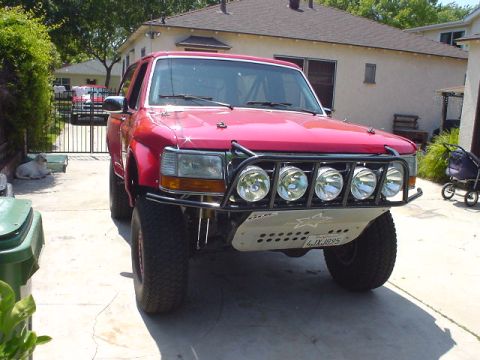 1984 Ford Bronco - This red SUV features a custom off-road bumper with multiple lights and a license plate reading 4JXJ895.