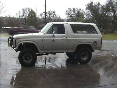 1984 Ford Bronco - A white Ford Bronco is parked on a wet surface with a "FOR SALE" sign in the window.
