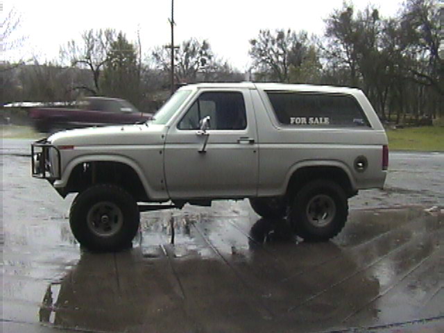 Lifted silver full-size Ford Bronco with For Sale lettering in the rear window, side view on wet pavement