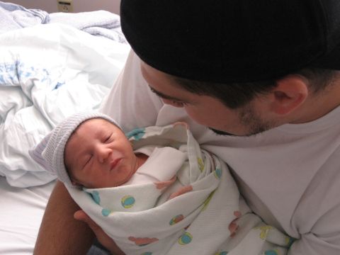 1990 Ford Bronco - A man in a black beanie and white t-shirt looks down at a swaddled newborn in a striped knit cap that he is holding on a hospital bed.