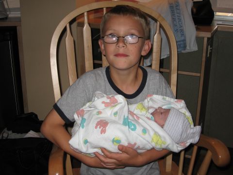 1990 Ford Bronco - A young boy in glasses sits in a wooden rocking chair in a hospital room holding a swaddled newborn wrapped in a printed receiving blanket.